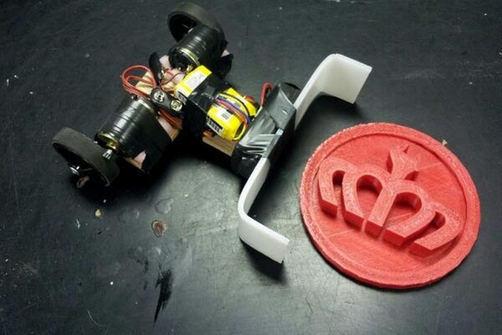 two wheeled remote control bot with a plow in front next to a 3D printed disk with the Hackerspace Charlotte logo