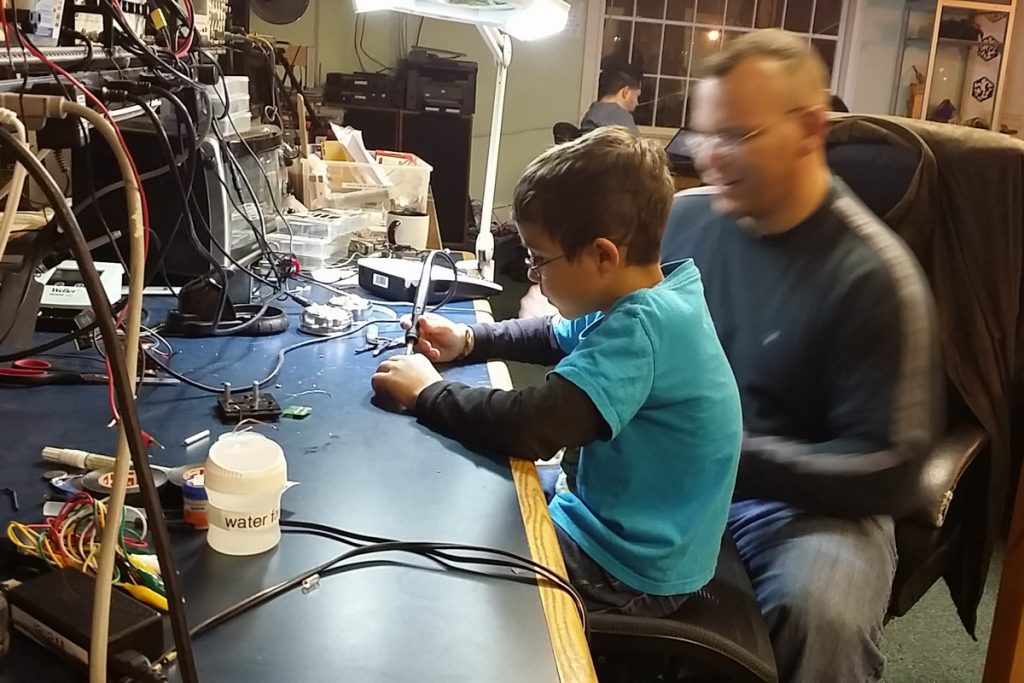 Father and son sit together practicing soldering at an electronics workbench