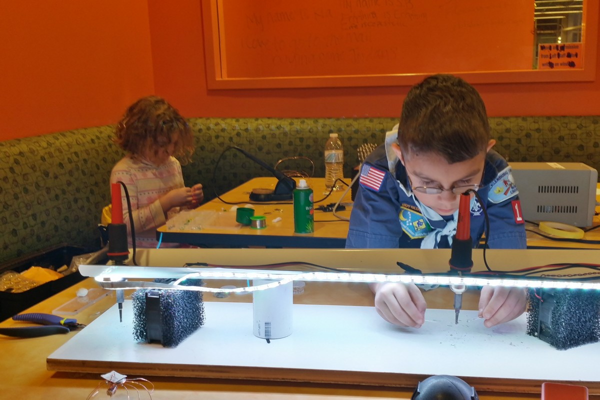 Kids soldering. One child has a traditional soldering iron, the other is at a station where the soldering iron is held in place and the components and solder are moved under the held iron.