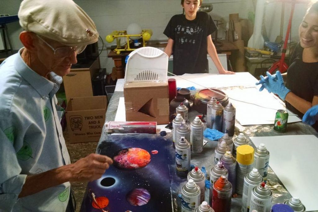 A table with lots of spray paint and an adult show kids how to make space paintings