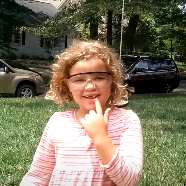 girl wearing safety goggles pointing at a loose tooth in her mouth